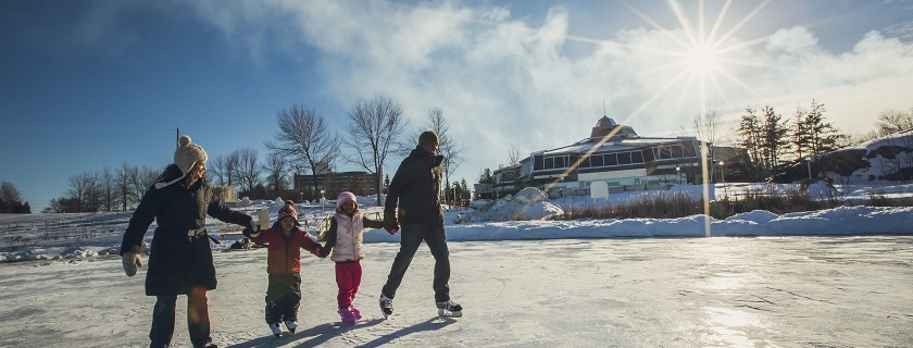 Two adults and two children holding hands skating on the Ramsey Lake skating path with Science North in the background.